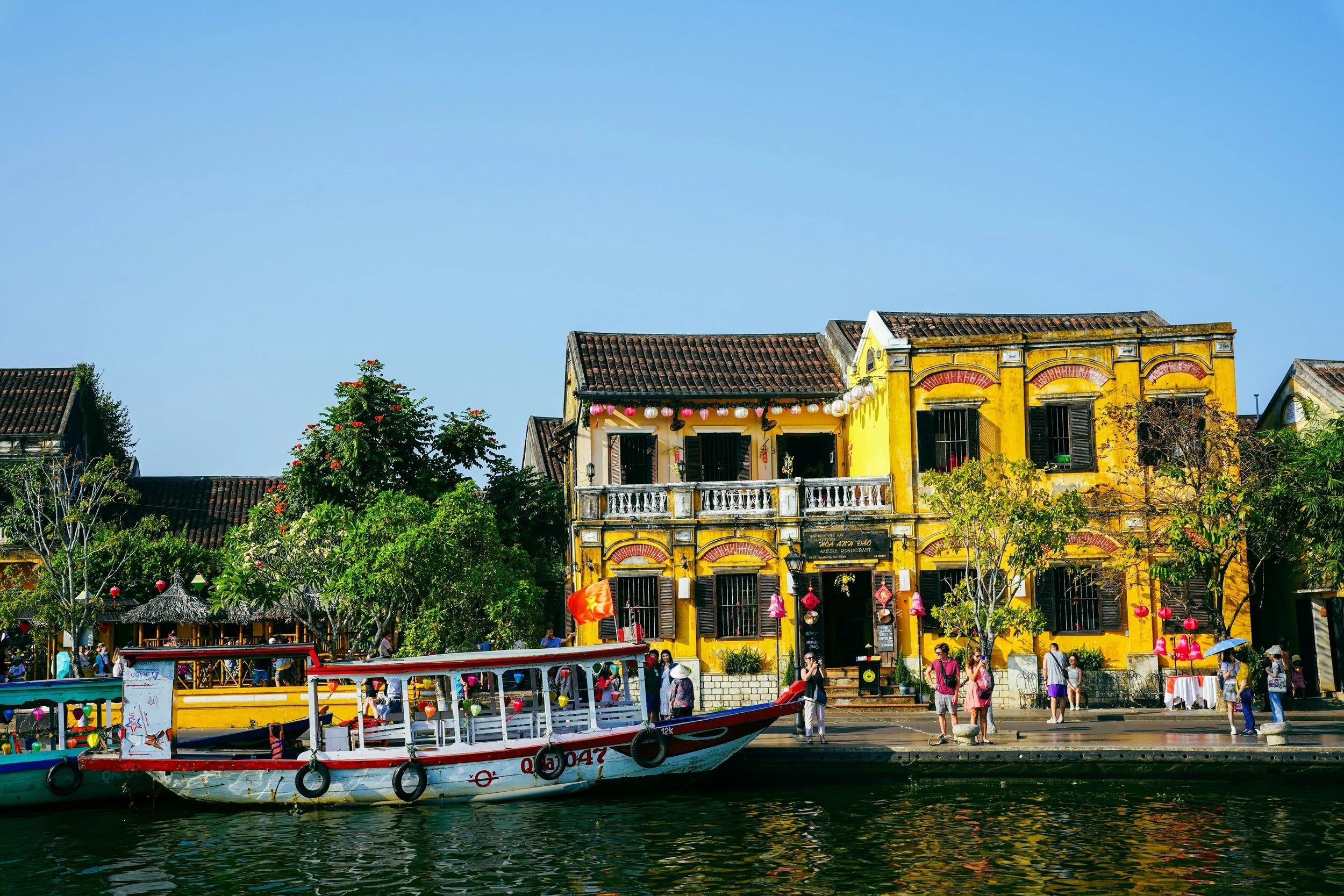 A vibrant yellow building with red and white lantern decorations and a balcony, situated along a river with boats and people on the riverbank.
