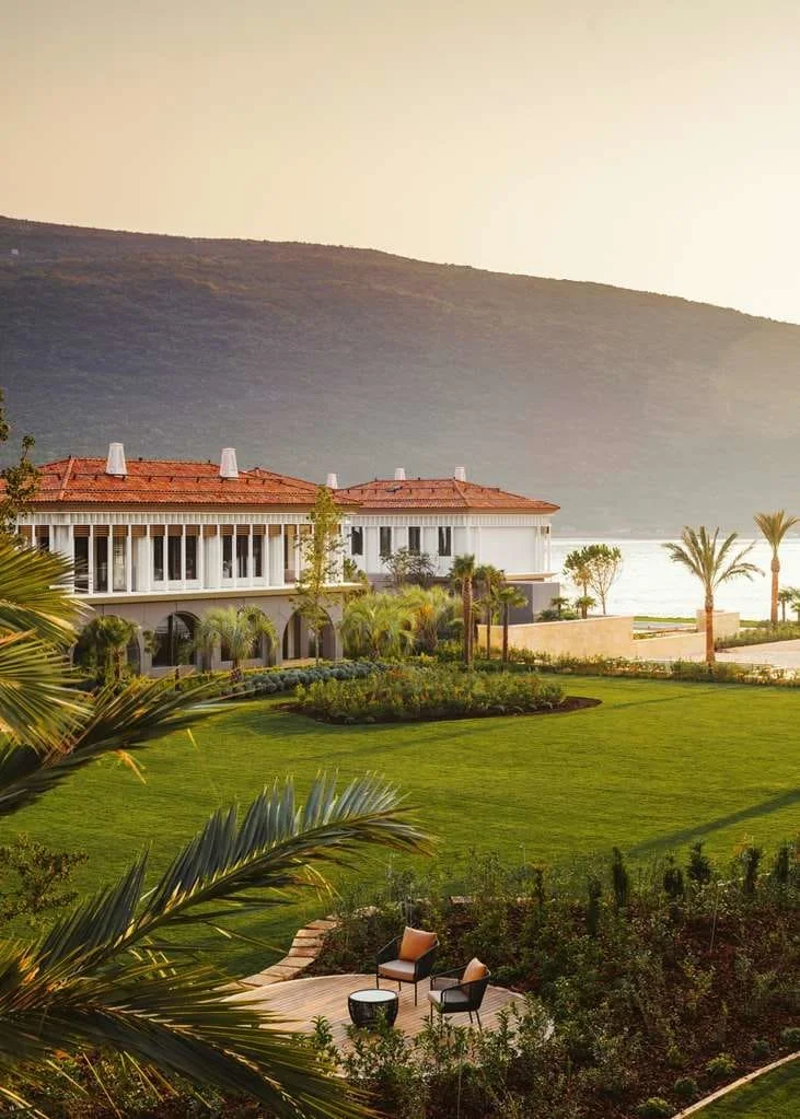 A luxurious house with a red-tile roof, large windows, and a white exterior, situated on a lush green lawn with palm trees, overlooking a body of water and mountains at sunset.
