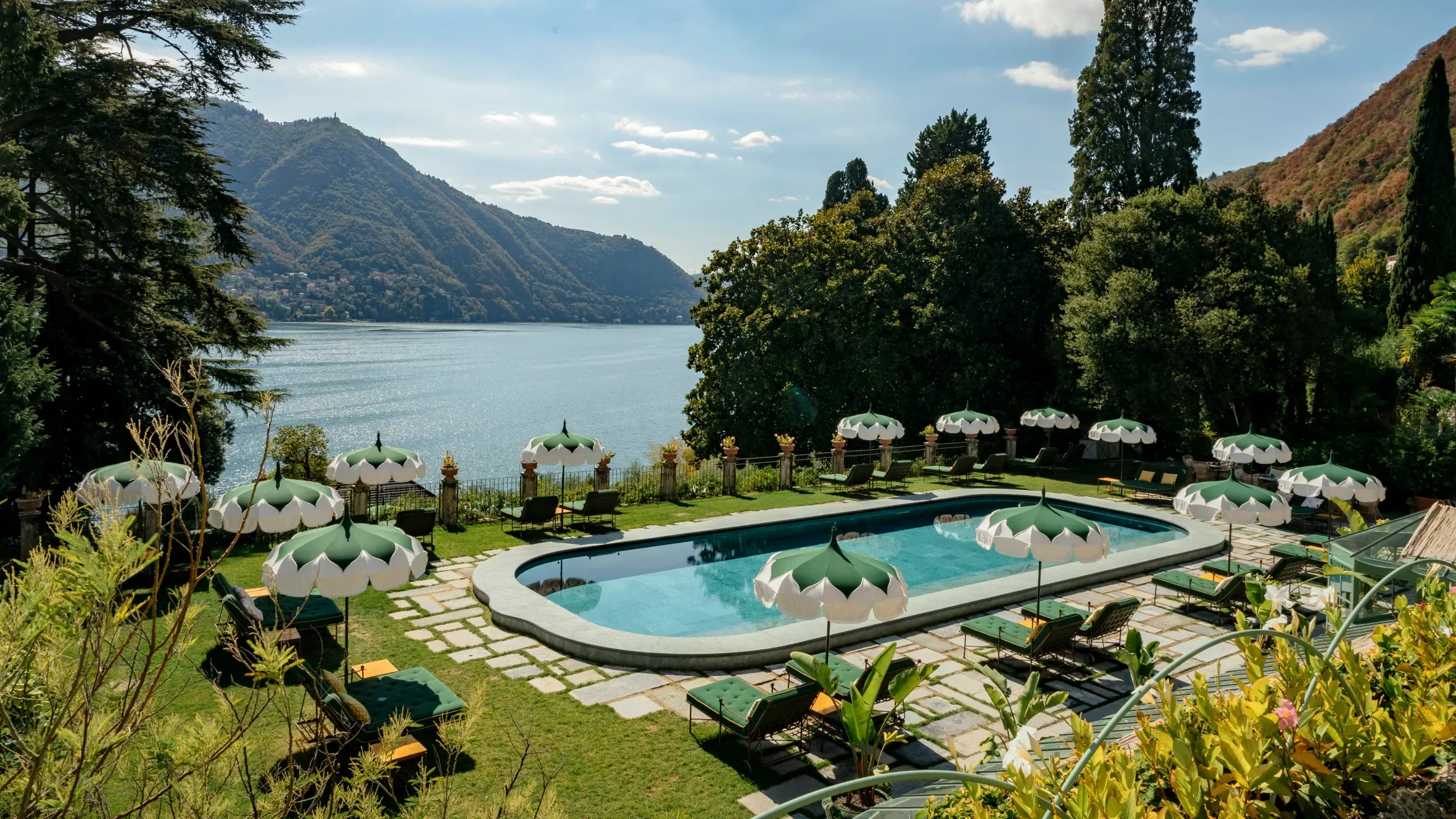 A scenic view of a swimming pool in a backyard overlooking a lake and mountains, with multiple umbrellas and lounge chairs around the pool.