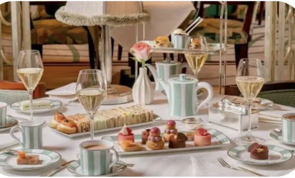A table set for a meal with plates, cups, glasses of white wine, a tiered serving tray with sweets, a plate of assorted desserts, a small vase with a pink flower, and teapots. The background includes chairs and a cozy setting.