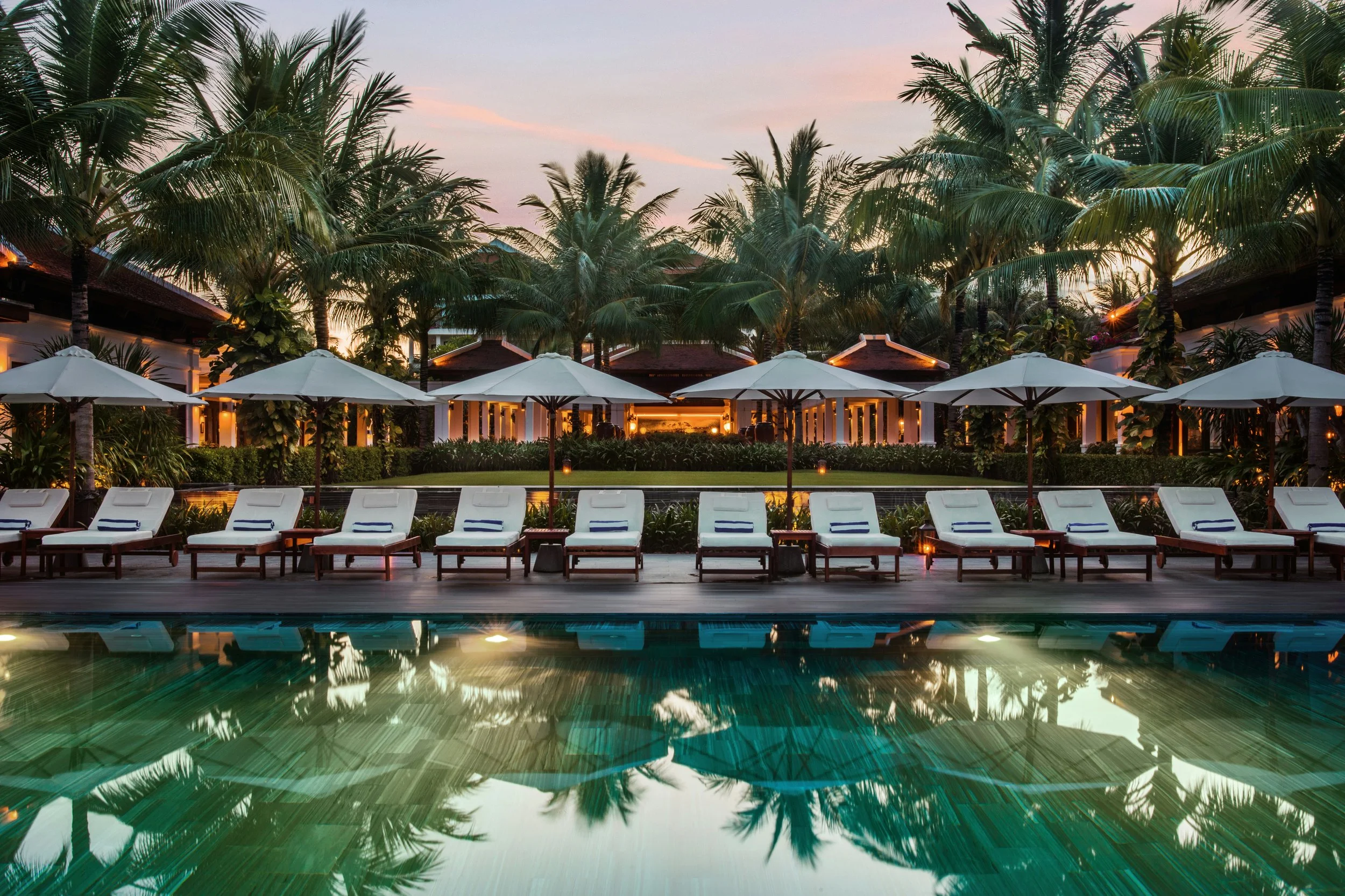 Poolside lounge chairs with white umbrellas at sunset, lush palm trees in the background, and resort buildings illuminated with warm lighting.