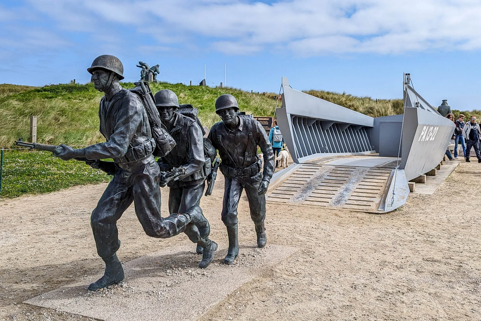 Bronze statues of soldiers running, set in an outdoor memorial site with grassy hills and a sign in the background, alongside a large military boat display and visitors walking nearby.