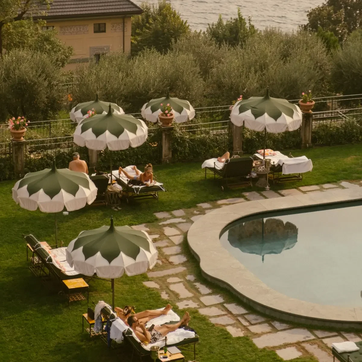 People relaxing by a swimming pool under large green and white umbrellas, some lying on lounge chairs, with a view of water and houses in the background.