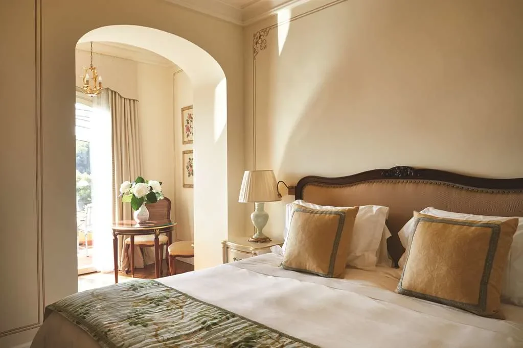 A bedroom with a large bed, decorative pillows, a nightstand with a lamp, and a small dining area with a table, two chairs, and a vase with flowers near a window with curtains.