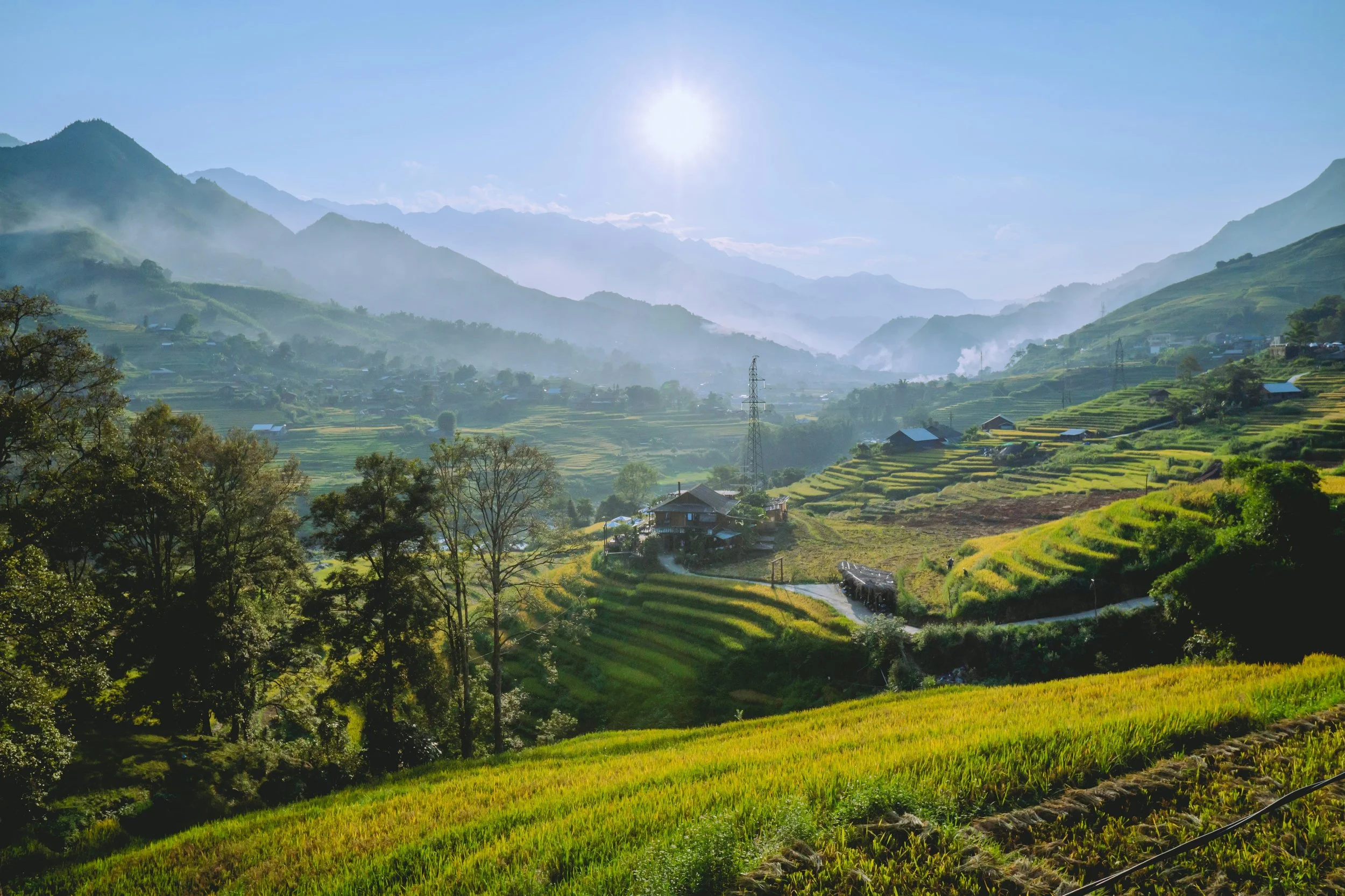 Sunrise over a valley with terraced rice fields, scattered houses, and rolling hills and mountains in the background.