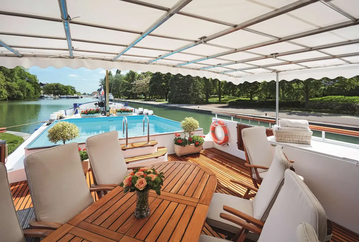 Luxury yacht deck with wooden table, white cushioned chairs, potted plants, and an inflatable pool, cruising along a tree-lined waterway under a white canopy.