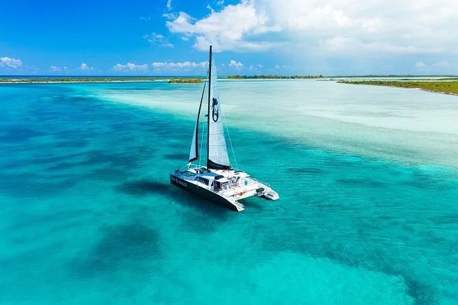 A sailboat on clear turquoise water near a sandy shoreline under a partly cloudy sky.