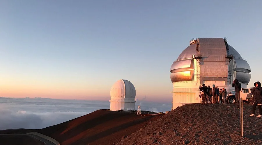 Observatories on a mountain at sunset, with a group of people and a vehicle nearby, under a partly cloudy sky.