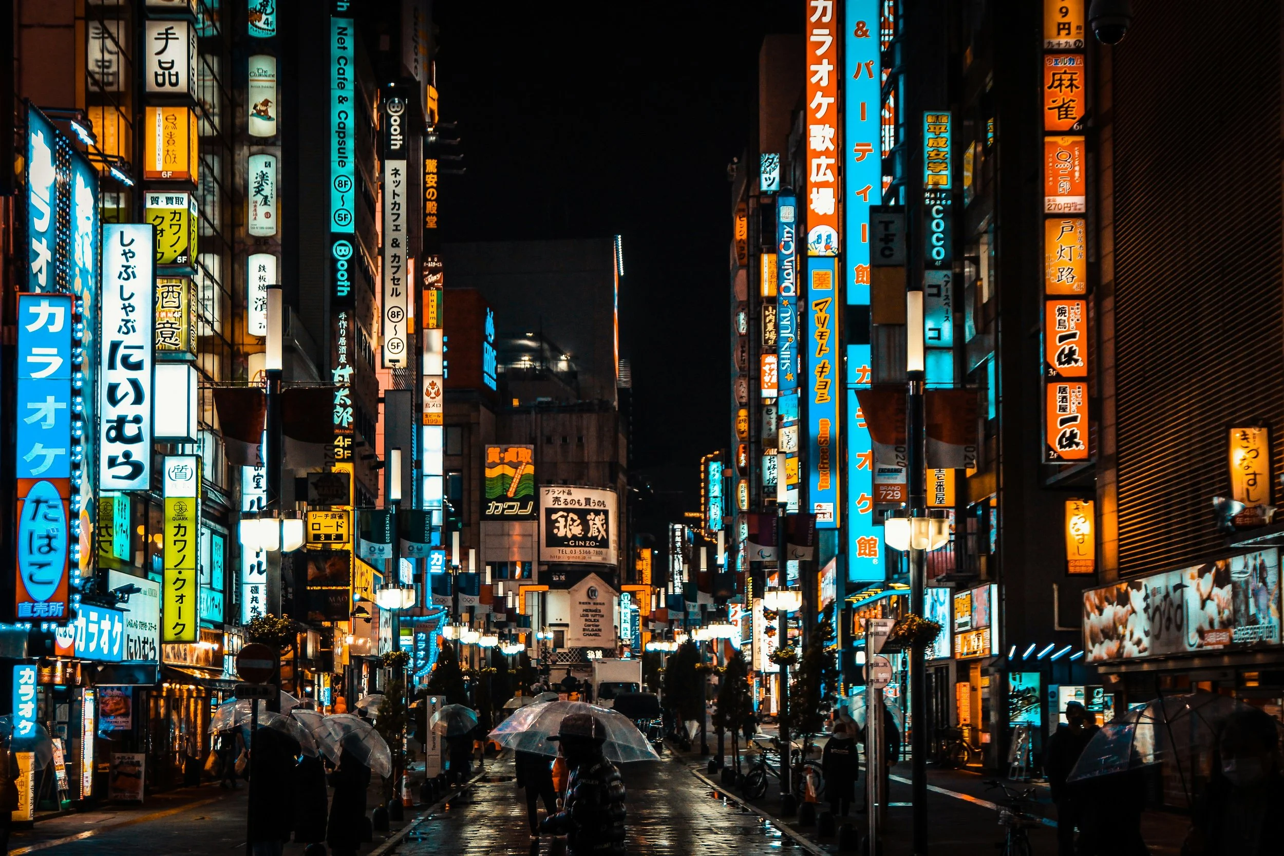 Nighttime city street scene illuminated with numerous bright, colorful neon signs in Japanese script. Pedestrians with umbrellas walk along the wet street amidst traffic and streetlights, creating a vibrant, lively atmosphere typical of an urban area in Japan.