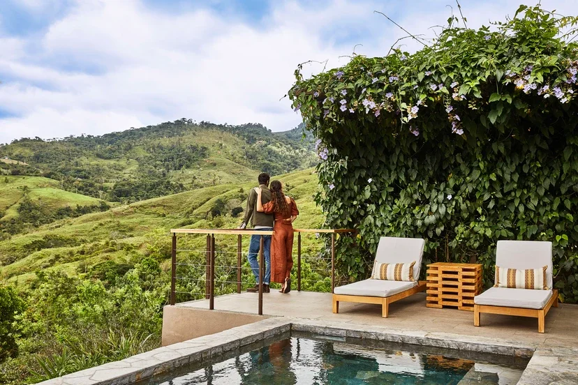 A couple standing on a balcony overlooking lush green hills, with two outdoor lounge chairs and a table on the patio.