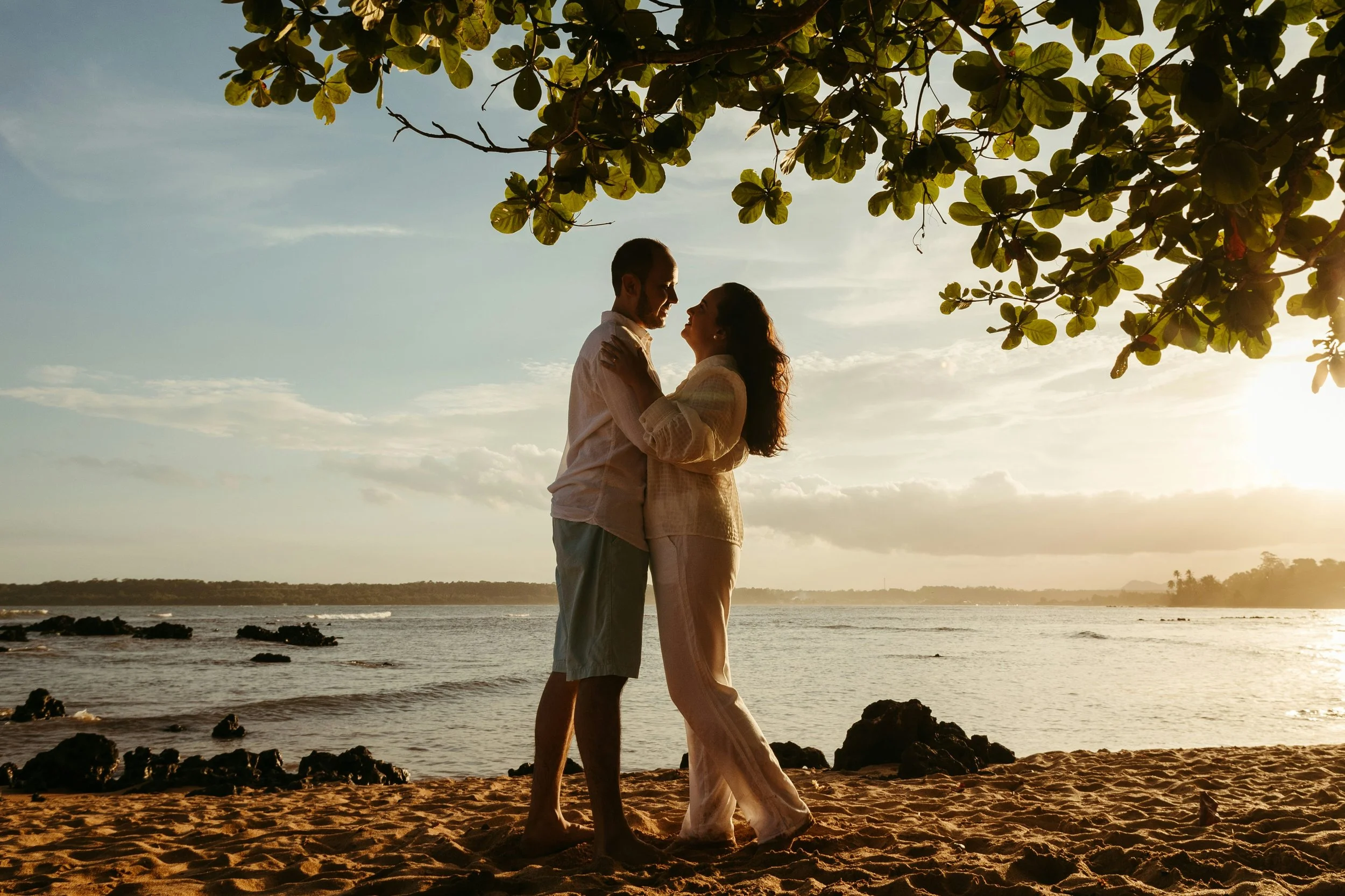A couple standing closely together on a beach during sunset, with the ocean and sky in the background and overhanging tree branches.