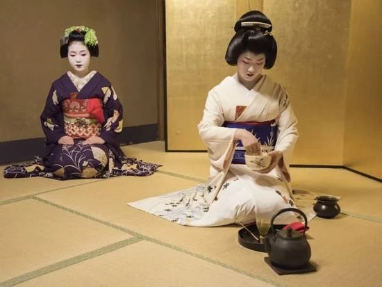 Two women dressed in traditional Japanese kimonos and elaborate hairstyles, sitting on tatami mats, with one preparing tea over a black kettle.