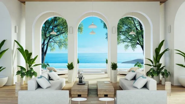 Bright living room with large arched windows overlooking the ocean, furnished with white sofas, potted plants, and wooden coffee tables.