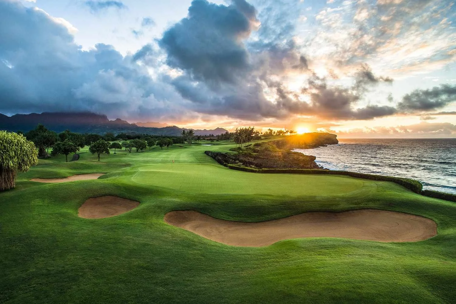 Sunset over an oceanfront golf course with green fairways, sand bunkers, and trees, with mountains in the distance and a partly cloudy sky.