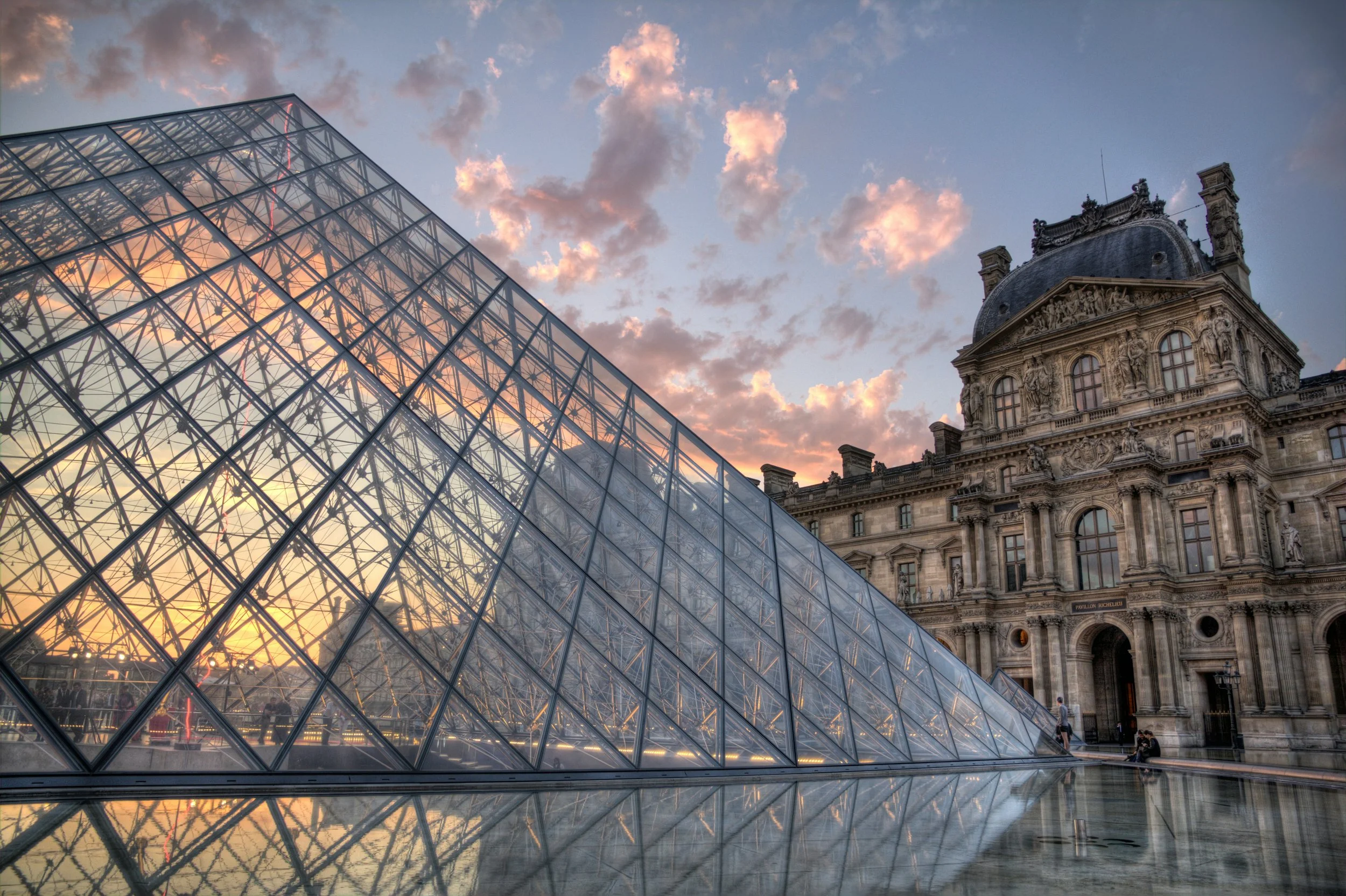 The image shows the Louvre Pyramid made of glass in front of the historic Louvre Palace in Paris, France. The sky is partly cloudy with pink and orange hues from the sunset.