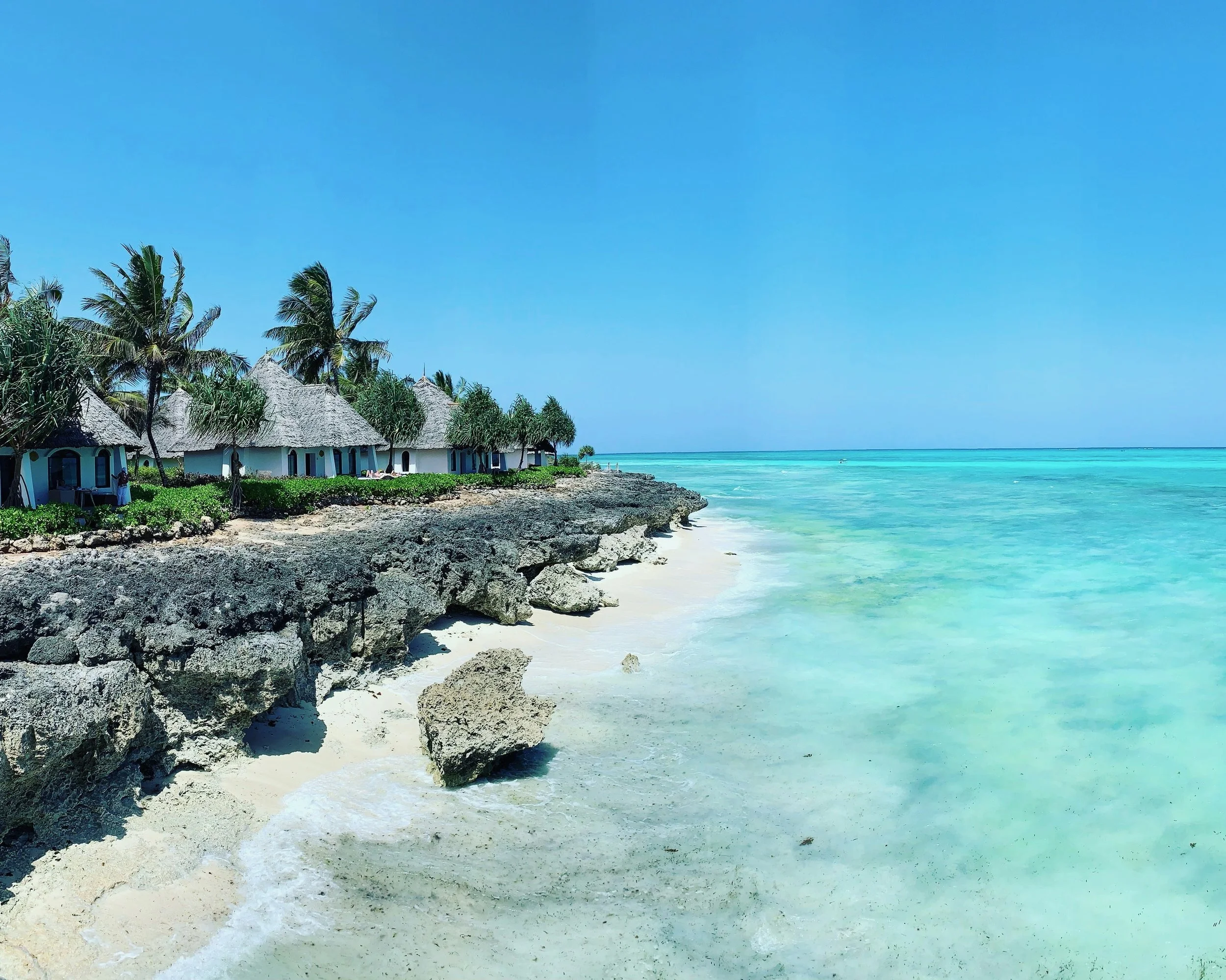 A tropical beach with thatched roof huts, palm trees, sandy shore, rocky edge, and turquoise ocean under a clear blue sky.