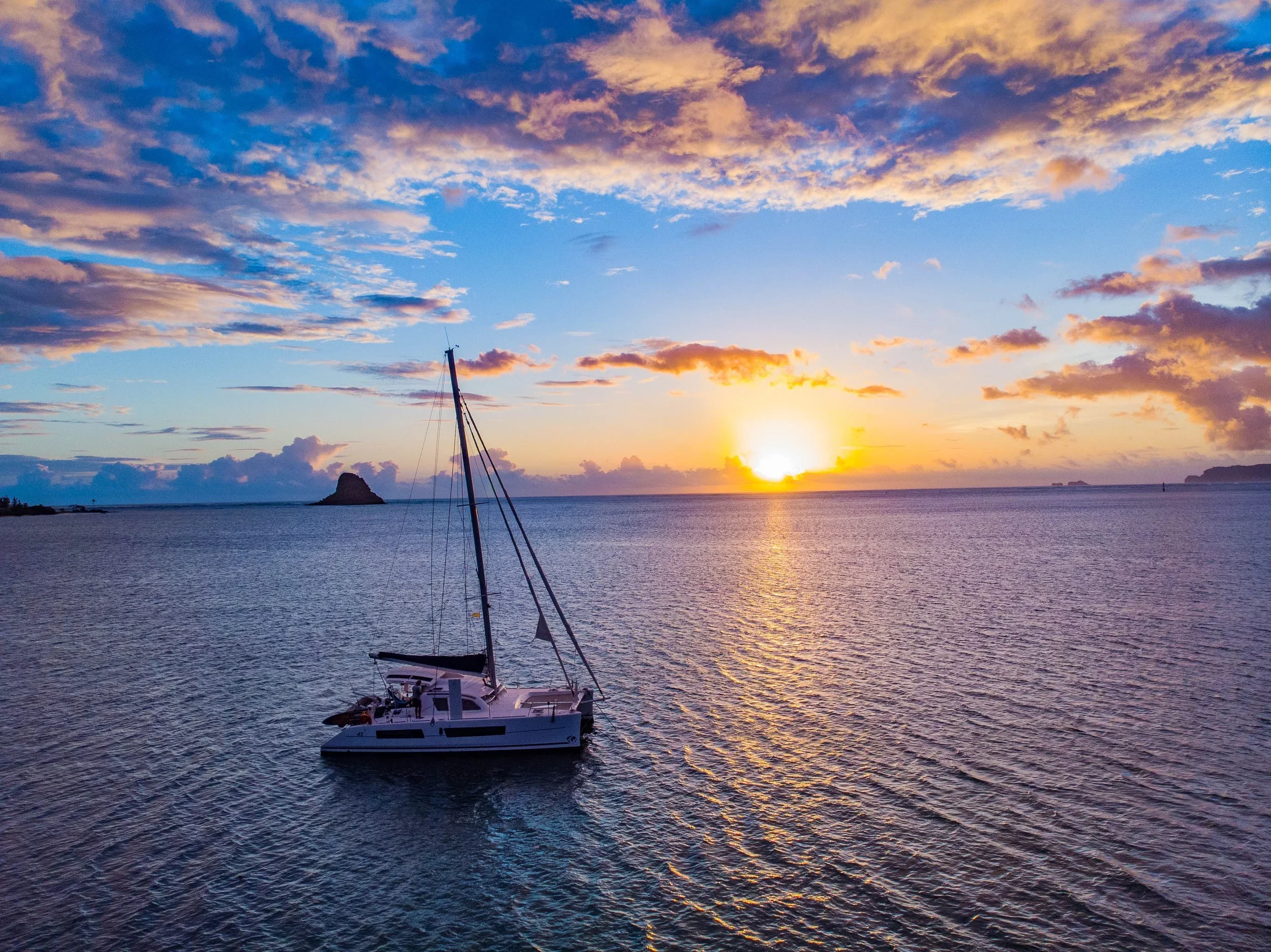 A sailboat floating on calm water during sunset with a vibrant sky filled with clouds in shades of purple, pink, and orange, and small islands in the distance.