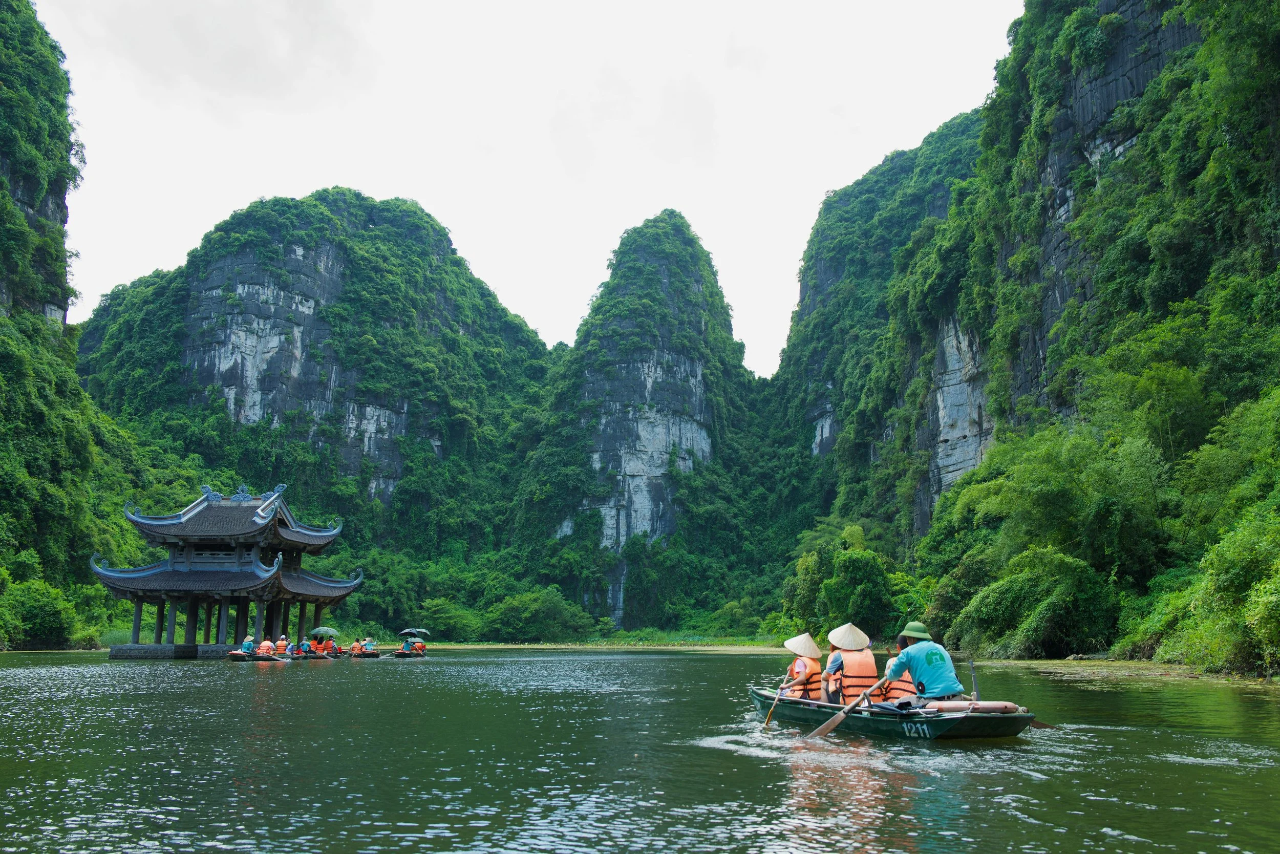 People in life jackets enjoying a boat ride on a river surrounded by lush green mountains and traditional pavilion with curved roof in a scenic landscape.