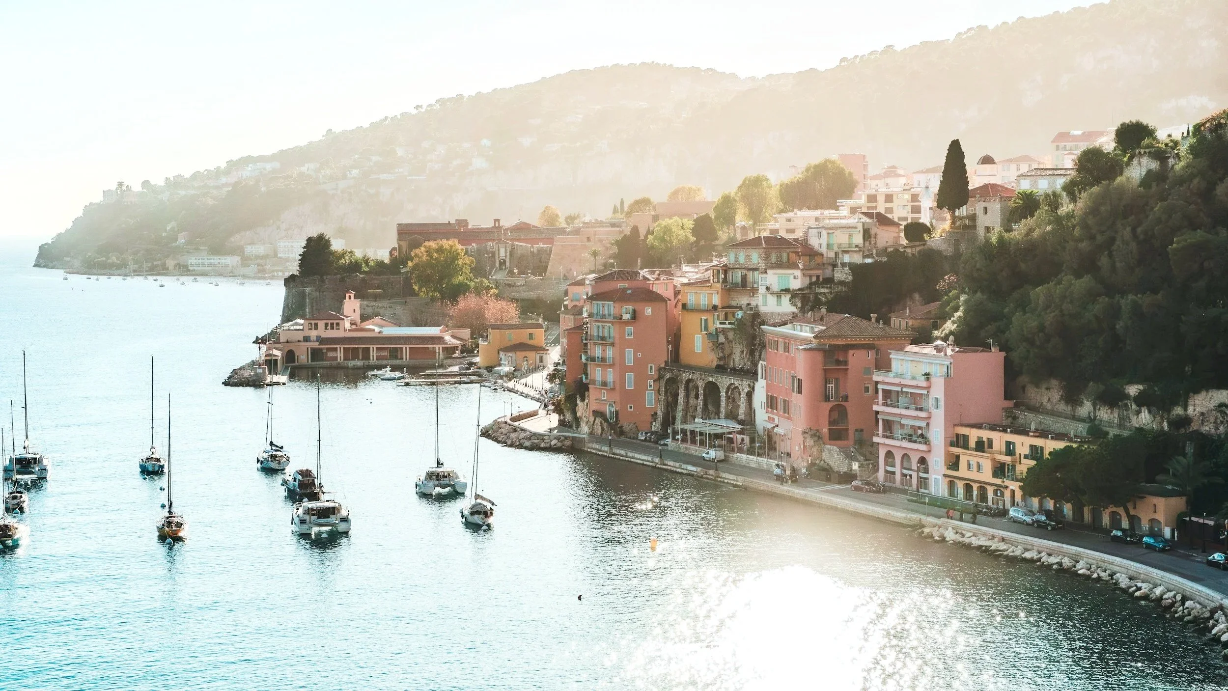 Colorful houses line the hillside next to a harbor with sailboats on the French Riviera. The scene is bathed in soft sunlight, creating a calm and scenic coastal view.