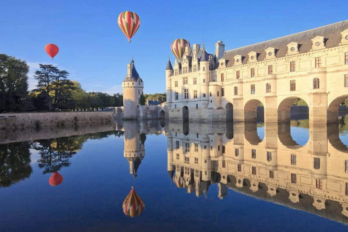 A castle reflection in a calm moat with three hot air balloons in the sky.