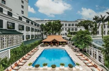 An outdoor hotel pool area with lounge chairs, surrounded by white buildings and palm trees.