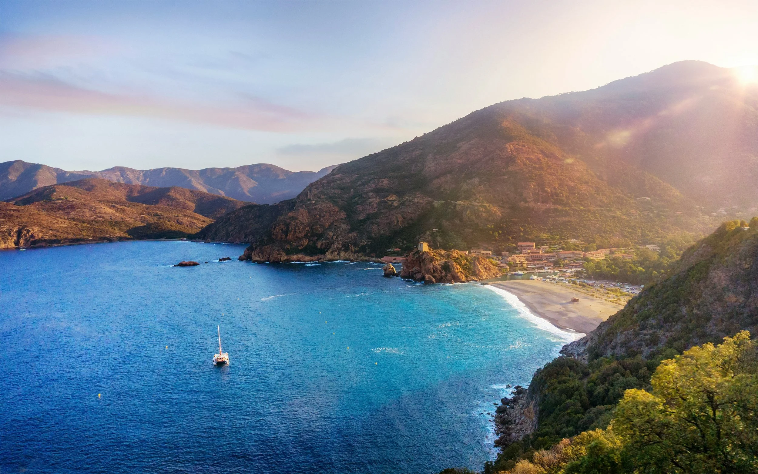 Scenic coastal view of a beach with mountains in the background, a sailboat on the blue water, and a small town at the foothills of the mountains during sunset.