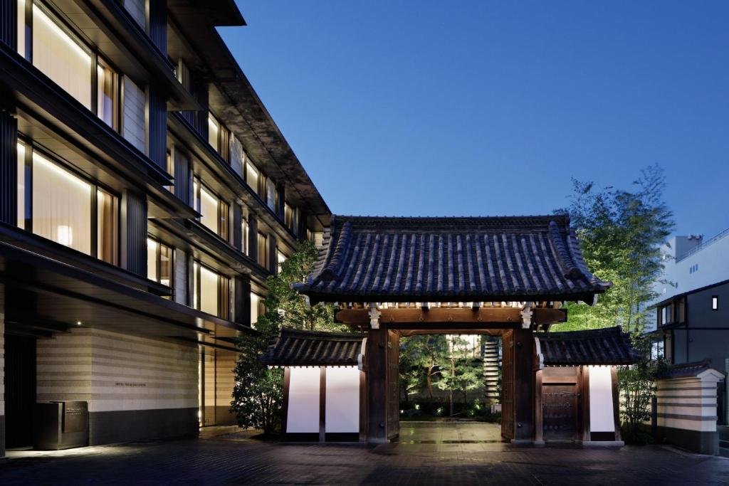 Traditional Japanese wooden gate with tiled roof in a courtyard at dusk, flanked by modern buildings and lush greenery.