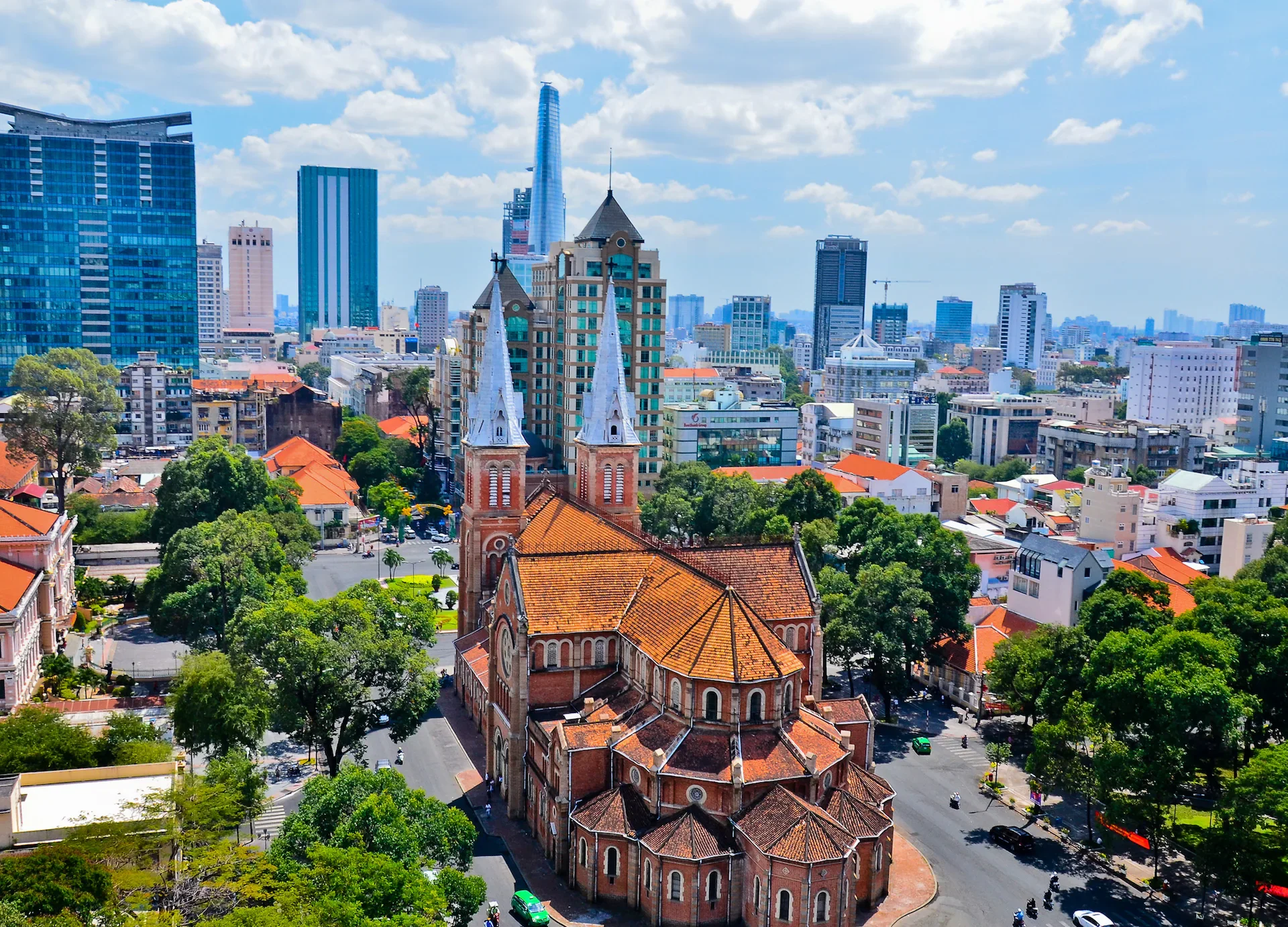 A cityscape featuring a historic red-brick church with two white spires in the foreground, surrounded by trees and modern high-rise buildings under a partly cloudy sky.