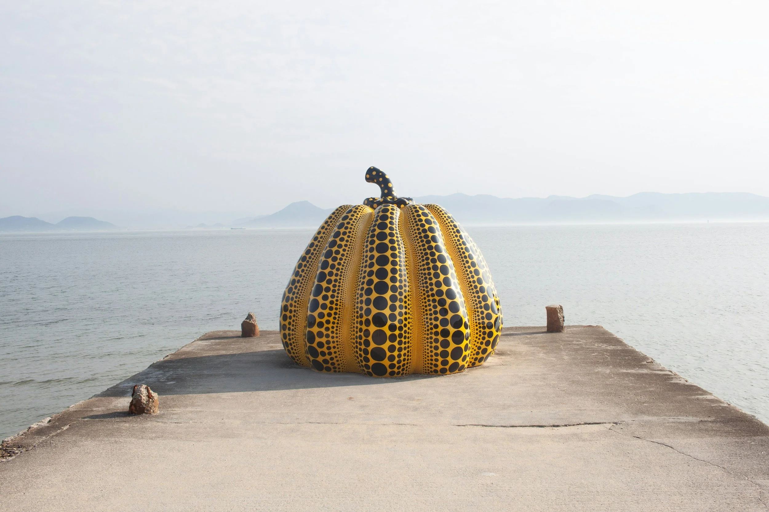 A large pumpkin sculpture painted yellow with black polka dots located on a concrete platform by a body of water, with mountains in the background under a cloudy sky.