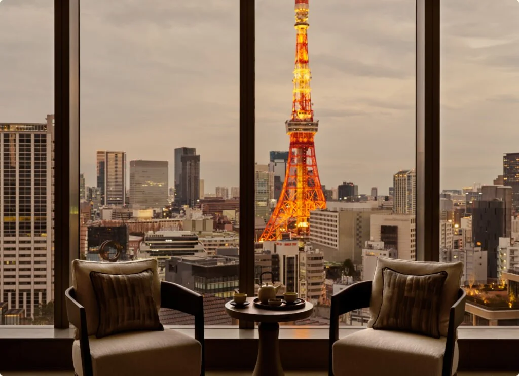 View from inside a room showing two chairs and a small table with tea set, overlooking Tokyo skyline with illuminated Tokyo Tower at dusk.