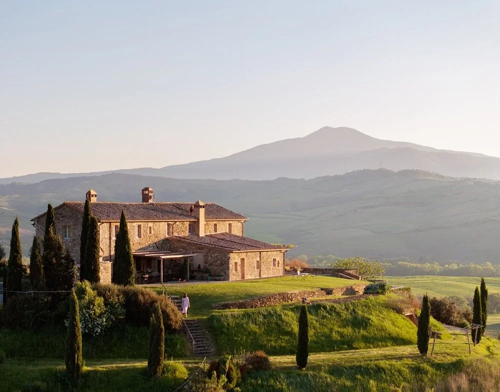 A stone farmhouse surrounded by tall cypress trees on a lush green hill with mountains in the background at sunset.