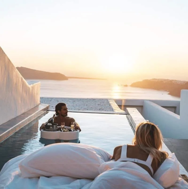 A woman lying in bed and looking at a man relaxing in a hot tub with a sunset view over the ocean.