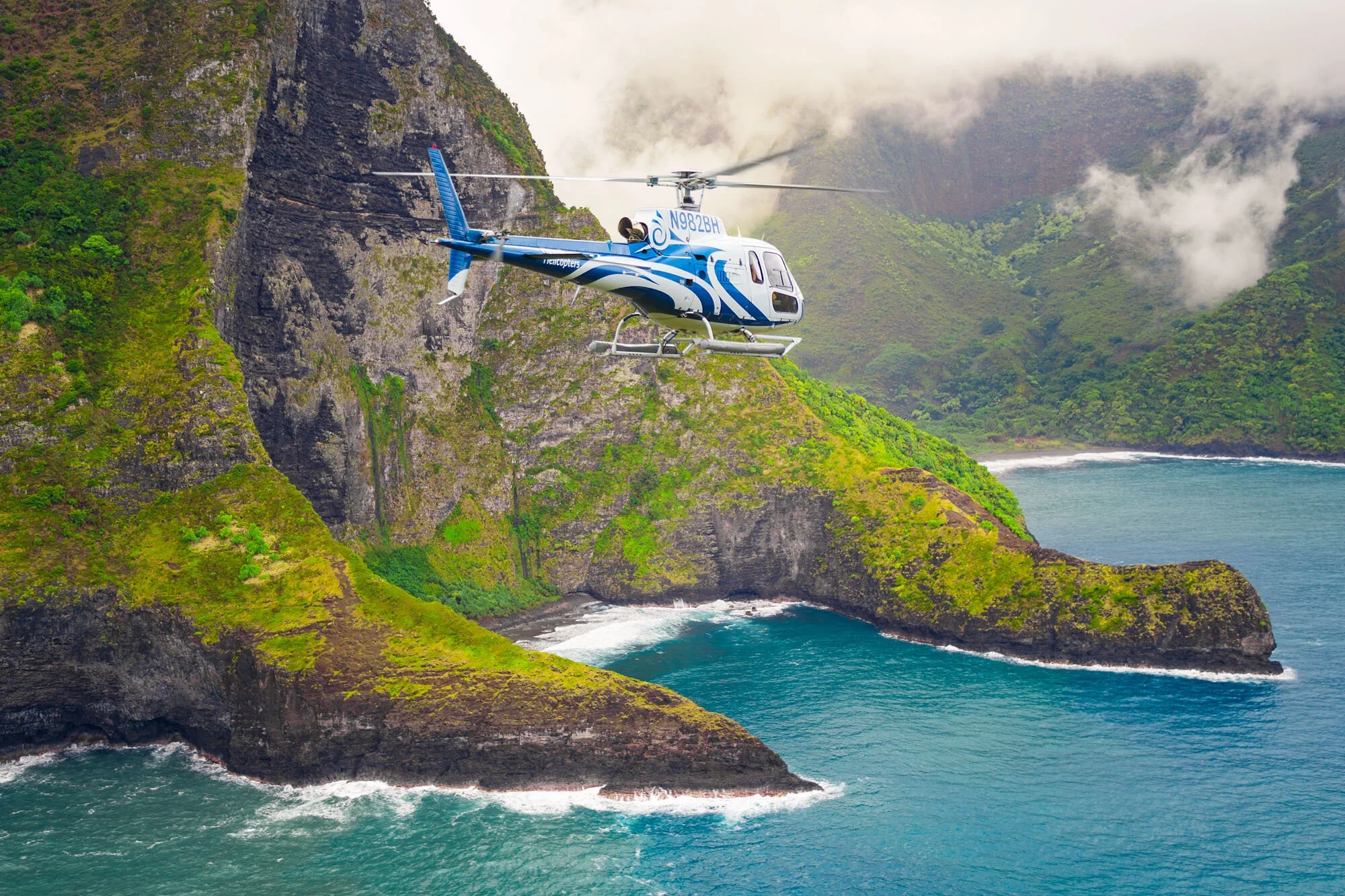 A helicopter flying near lush green cliffs over the ocean with rocky formations and waves.