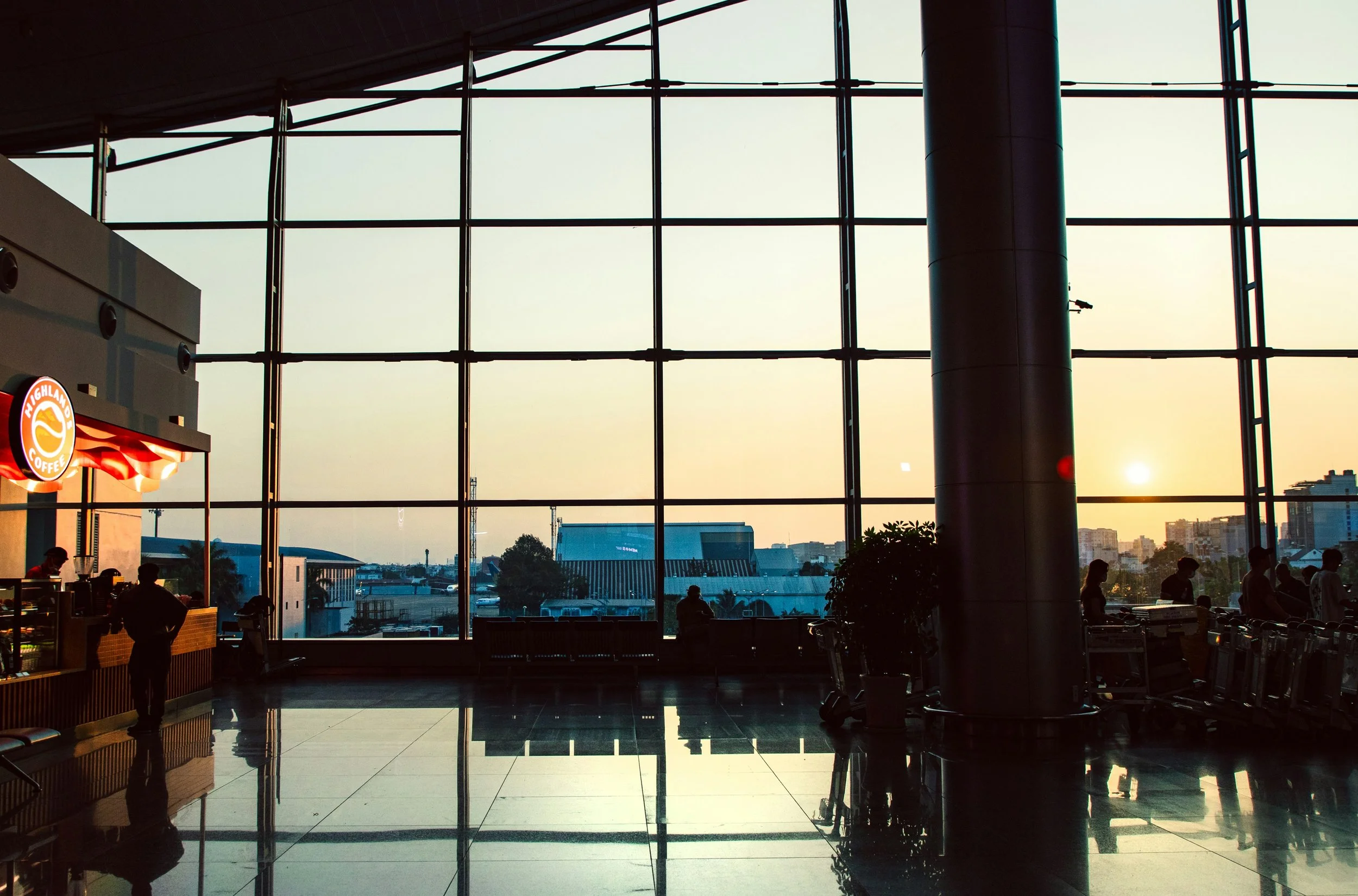 Interior of an airport terminal with large glass windows showing an urban skyline at sunset, people in silhouette, and a Highland Coffee store on the left.