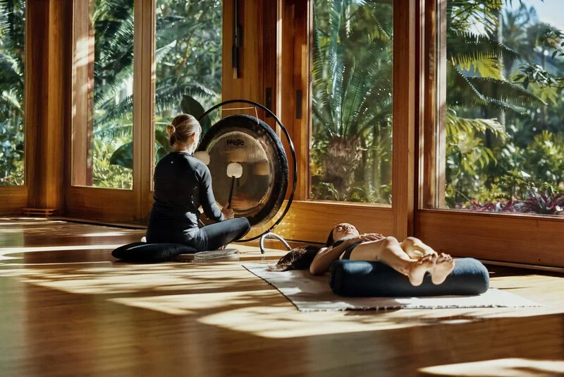 A woman sitting on a cushion playing a gong while another woman lies on a cushion relaxing nearby in a sunlit room with large glass windows and a view of lush green tropical plants outside.