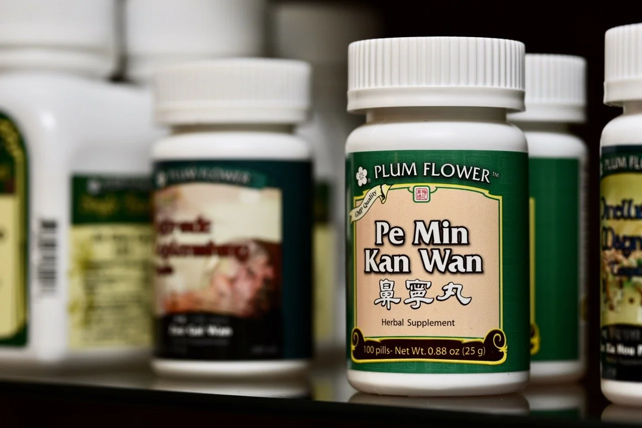 A bottle labeled 'Pe Min Kan Wan' herbal supplement on a store shelf, with other supplement bottles blurred in the background.