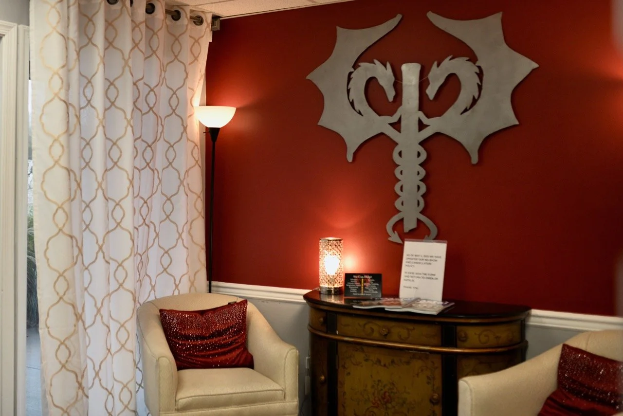 A cozy room corner with a deep red wall, a decorative metallic wall art piece, two beige armchairs with red cushions, a wooden console table, a tall floor lamp, and a sheer curtain with a gold pattern covering a window.