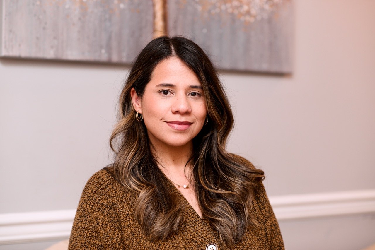 A woman with long wavy brown hair wearing gold earrings and a brown sweater, sitting in front of a neutral-colored wall with artwork.