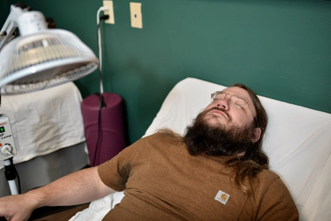 A man with long hair and a beard wearing glasses and a brown t-shirt is lying in an antigravity chair with his eyes closed.