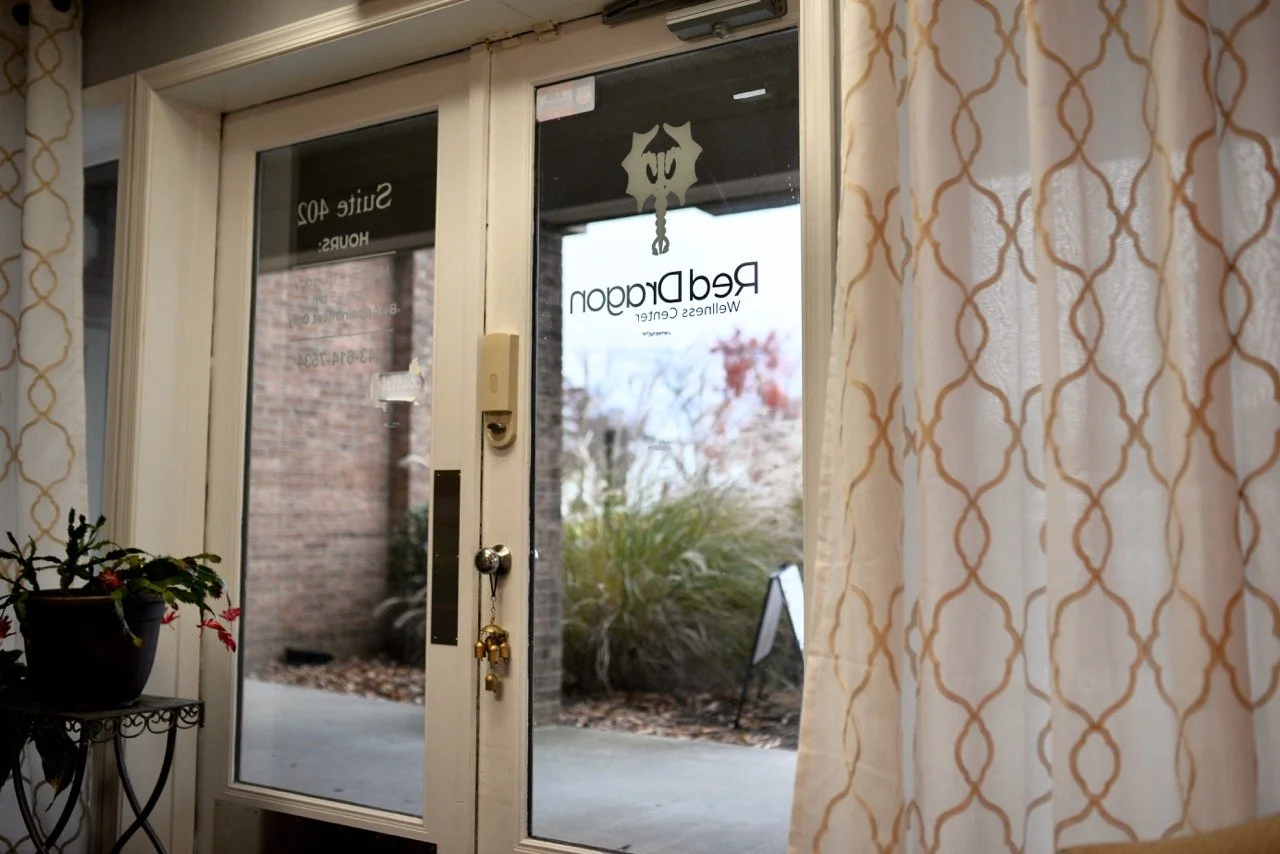 View through a glass door with a sign reading "Red Dragon Wellness Center" and keychains hanging from the handle. Curtains with a geometric pattern partially cover the view, and there is a potted plant on a small table near the door.