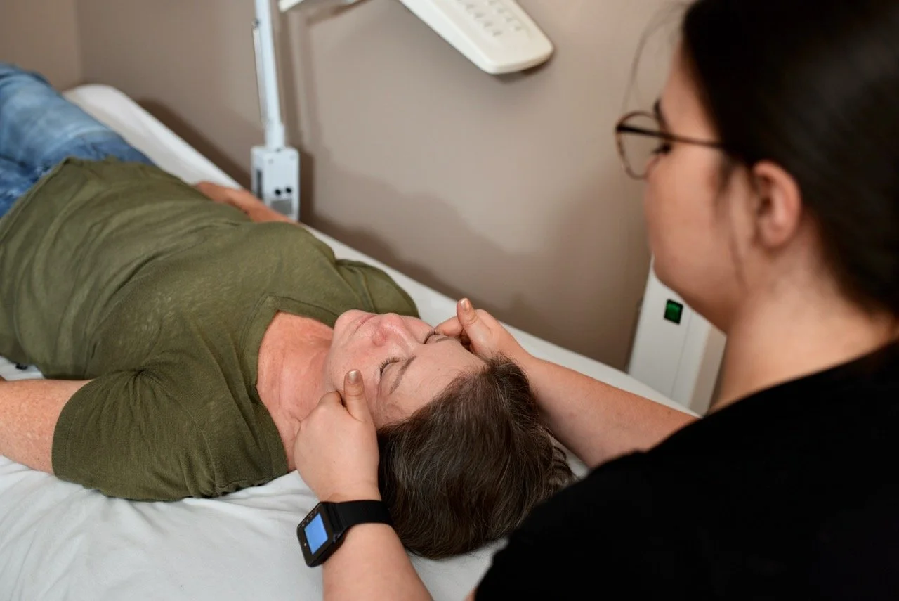 A woman lying on a hospital bed with her eyes closed, receiving a facial massage or therapy from another woman who is wearing glasses and a smartwatch.