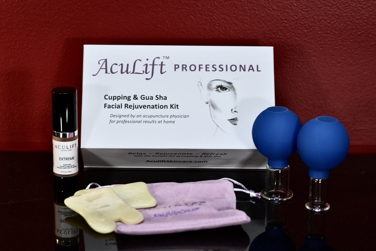 AcuLift facial rejuvenation kit with a box, two blue silicone cups, a small bottle of AcuLift Extreme serum, a purple cloth pouch, and a pair of yellow gloves on a black surface against a red wall.