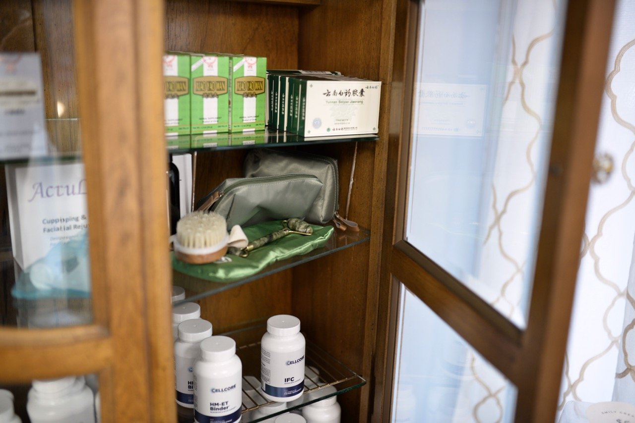 A wooden display cabinet containing various medical supplies, including medication bottles, a silver bag, herbal sticks, boxed health products, a face mask, and medical tools, situated next to a window with patterned curtains.
