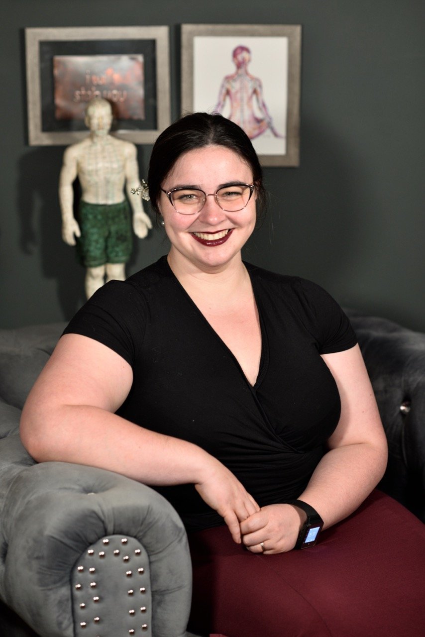 A young woman with dark hair, glasses, and dark lipstick, smiling and sitting on a gray tufted sofa in a room with dark green walls. Behind her, there are two framed art pieces and a doll-like figure in green shorts.
