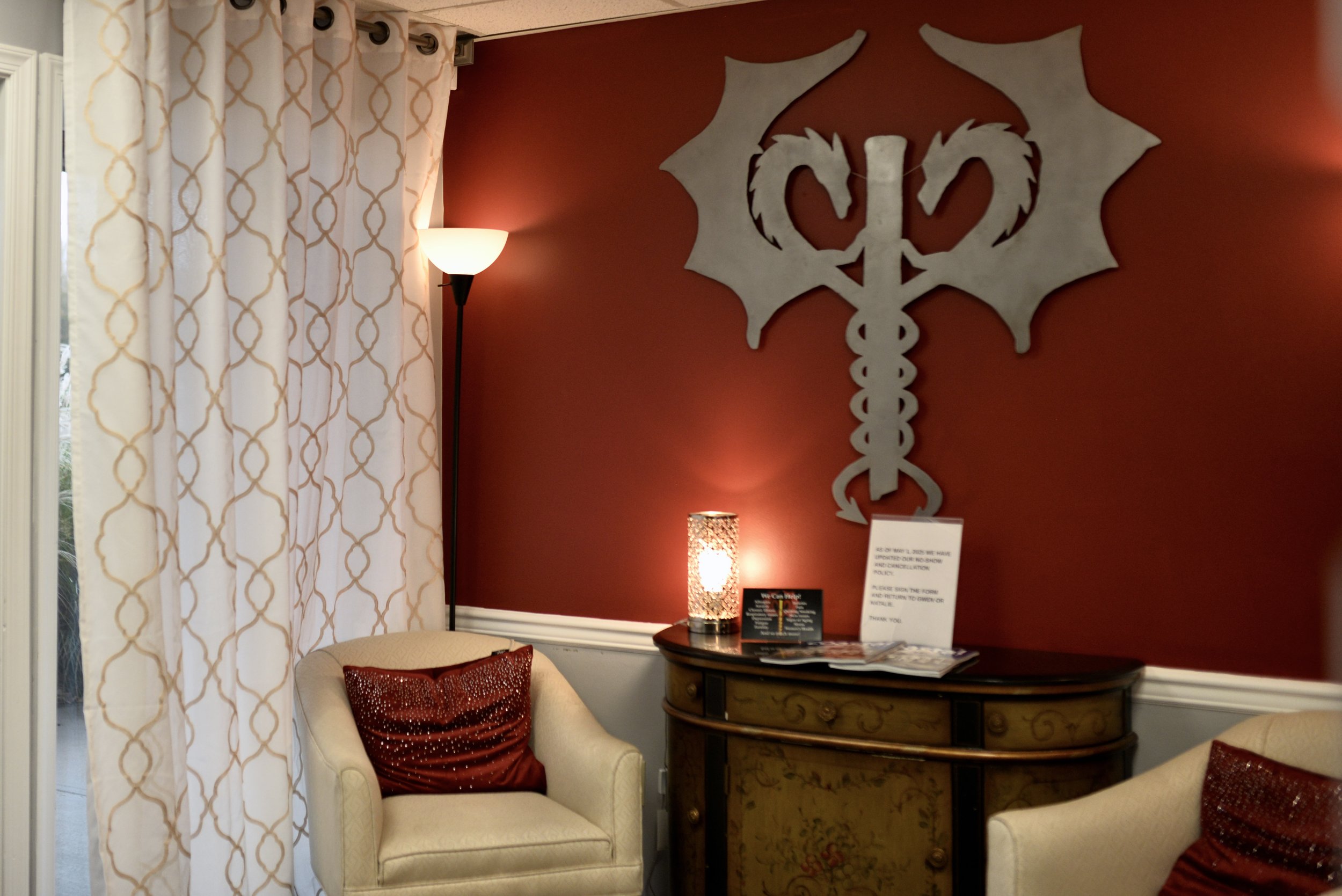 A cozy living room corner with two beige armchairs, each with a red sequin throw pillow, a curved wooden sideboard with decorative details, a tall lamp with a patterned shade, a standing floor lamp with a white shade, a red accent wall with a large medieval-style white dragon cross symbol, and a window with white curtains with gold geometric patterns.
