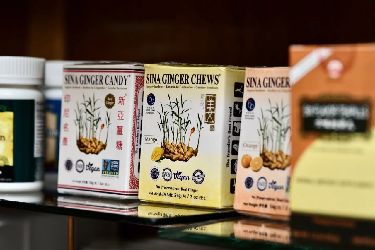 Boxes of Sina Ginger Chews in different flavors on a store shelf, featuring illustrations of ginger plants and labels indicating flavors like mango and orange.