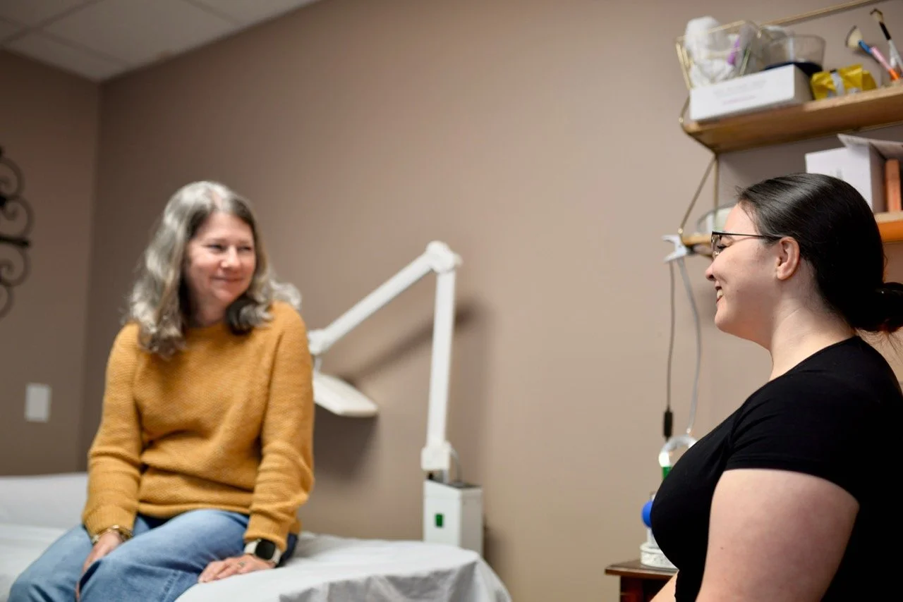 Two women having a conversation in a medical examination room. One woman is sitting on an examination table, wearing a mustard sweater and jeans, with a smartwatch on her wrist. The other woman, wearing glasses and a black shirt, is facing her, smiling. In the background, there is medical equipment and shelves with supplies.
