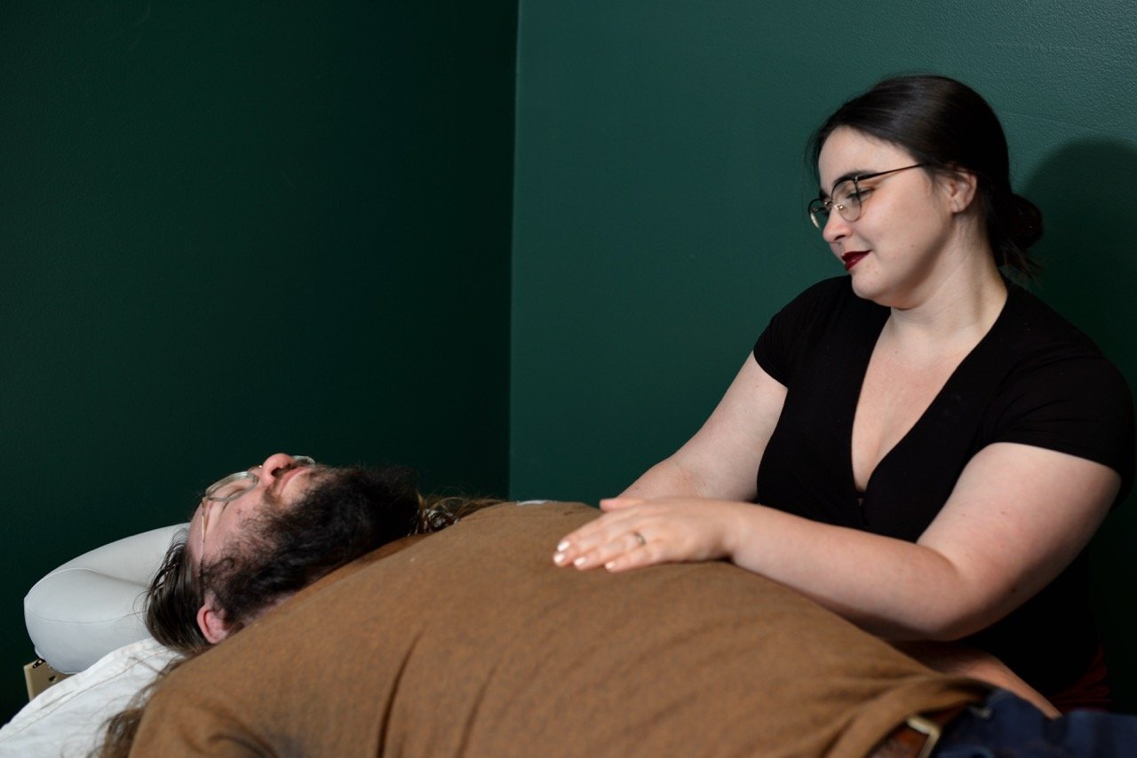 A woman with glasses smiling gently while resting her hand on a man's chest during a nervous system regulation session in a room with dark green walls.