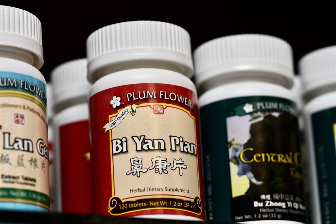 Bottles of herbal dietary supplements on a shelf, prominently featuring a red bottle labeled 'Bi Yan Pian' with Chinese characters, from the Plum Flower brand.