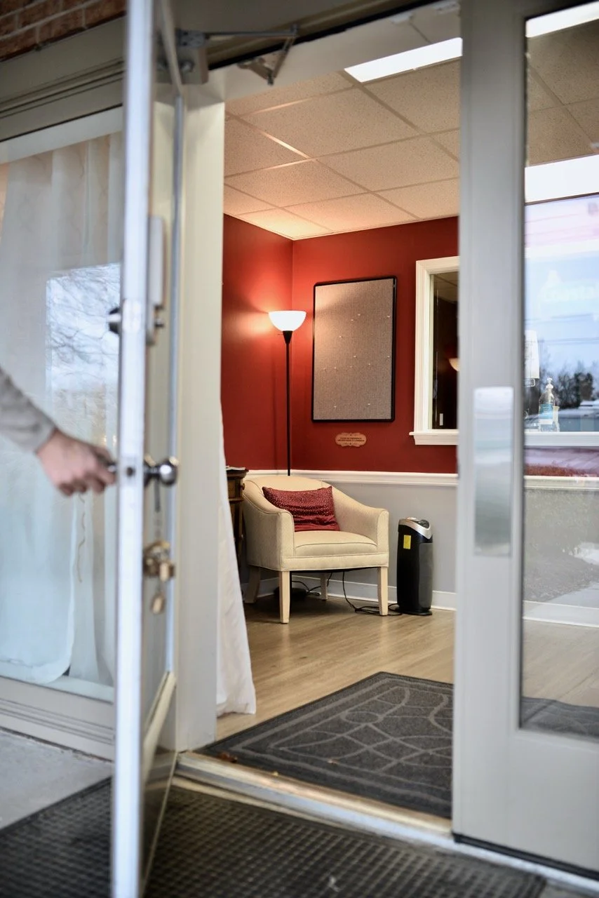 View through glass doorway into a cozy room with a beige armchair, a red pillow, a tall standing lamp, a framed bulletin board, and a window with outside view.